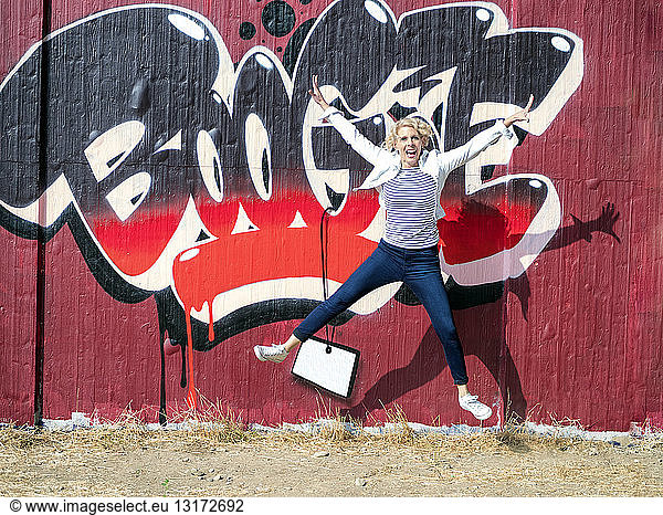 Happy mature woman jumping in the air in front of graffiti wall
