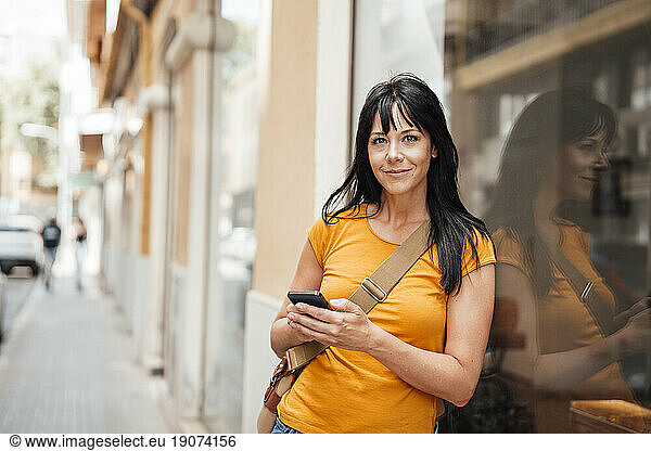 Happy mature woman leaning on glass with smart phone
