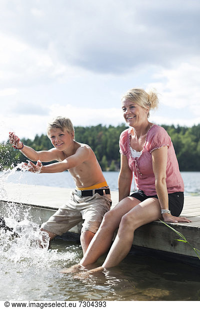 Happy mature woman sitting on pier with son splashing water