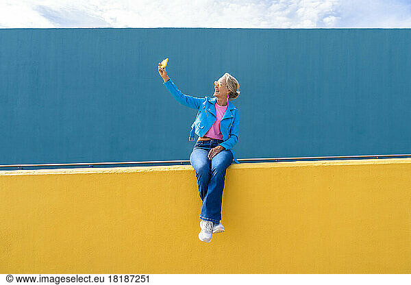 Happy mature woman taking selfie through mobile phone on wall