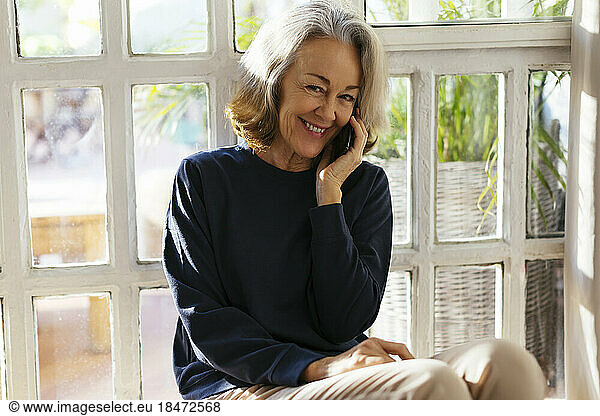Happy mature woman talking on smart phone in front of window
