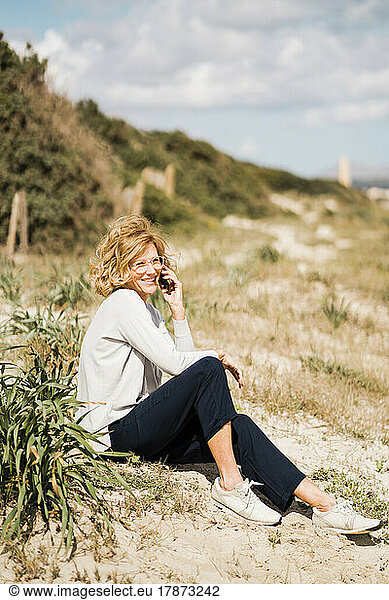 Happy mature woman talking on smart phone sitting by plant at beach