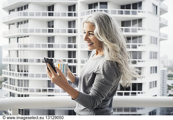 Happy mature woman using smart phone while holding coffee mug on balcony against building