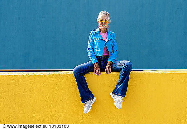 Happy mature woman wearing blue leather jacket on yellow wall