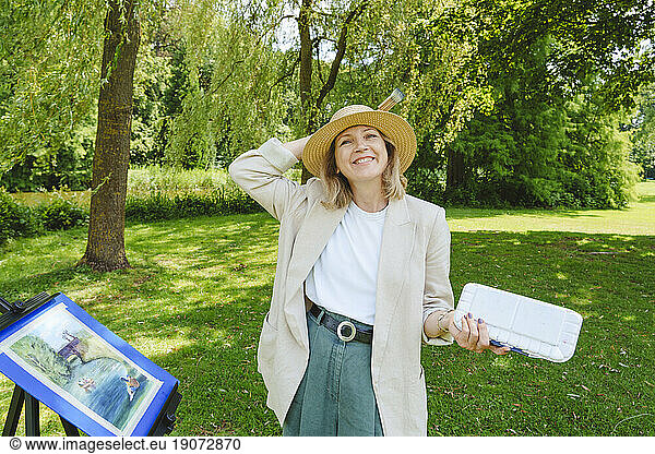 Happy mature woman wearing hat standing by painting in park