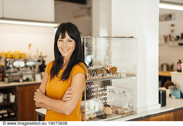 Happy mature woman with arms crossed standing by retail display in cafe
