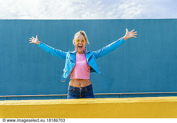 Happy mature woman with arms outstretched in front of blue wall
