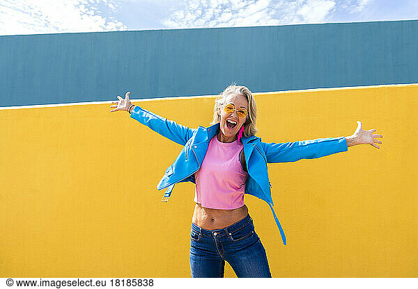 Happy mature woman with arms outstretched in front of colorful wall