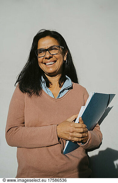 Happy mature woman with book and file standing against gray wall at university on sunny day