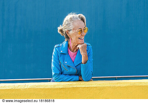 Happy mature woman with hand on chin in front of blue wall