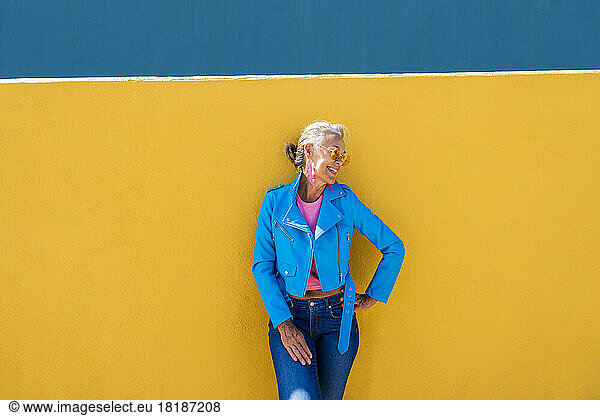 Happy mature woman with hand on hip in front of colored wall
