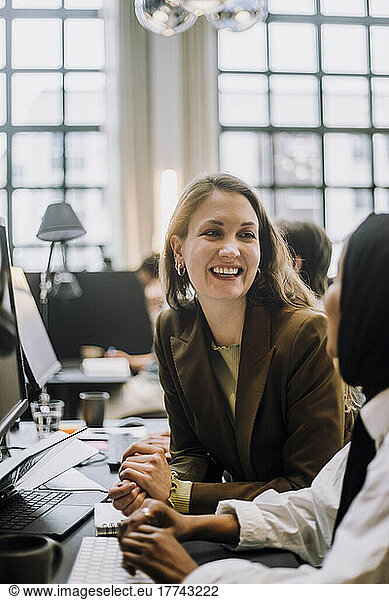 Happy mid adult businesswoman discussing with female colleague sitting at desk in office