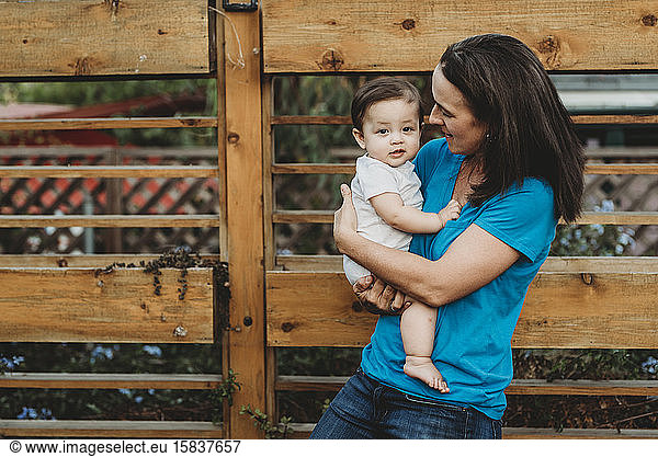 Happy mom holding smiling baby daughter outdoors near wooden fence