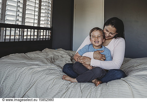 Happy mom holding smiling son on lap in bedroom near window
