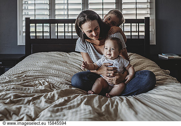 Happy mom on bed with baby on lap and son hugging her from behind