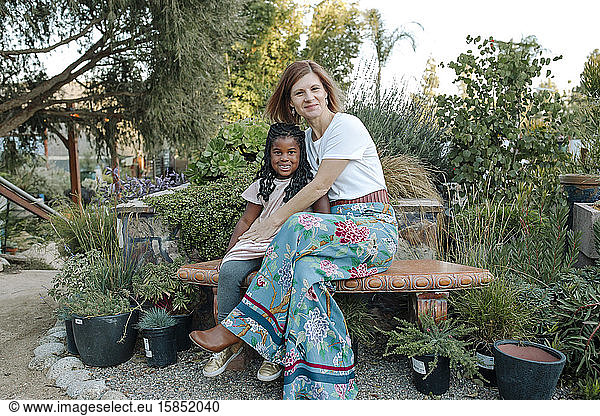 Happy mom & smiling black daughter on bench in front of lush plants