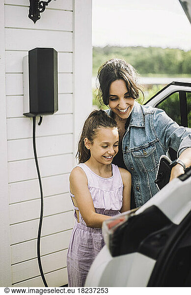 Happy mother and daughter charging electric car together near house