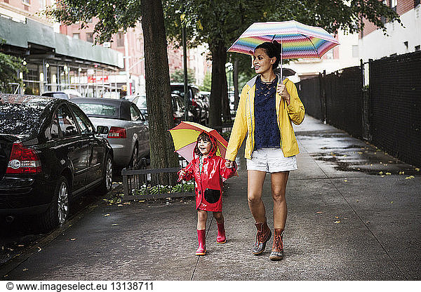 Happy mother and daughter holding umbrellas while walking on sidewalk