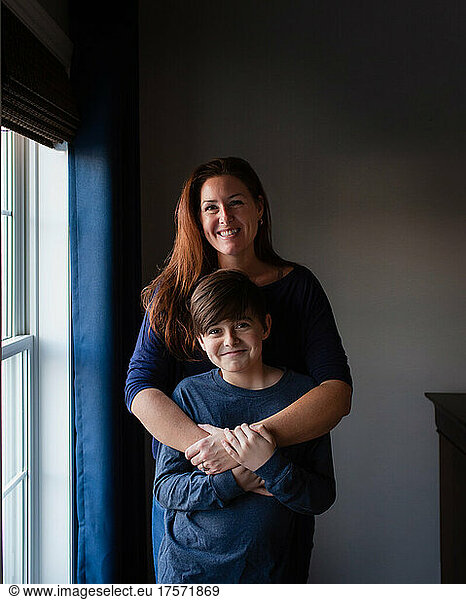 Happy mother and son embracing and smiling indoors beside a window.