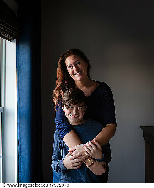 Happy mother and son embracing and smiling indoors beside a window.