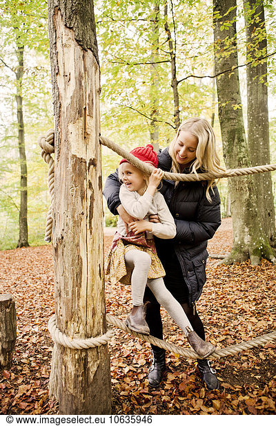 Happy mother assisting daughter balancing on rope in forest