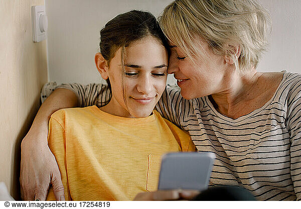 Happy mother embracing daughter using mobile phone in bedroom