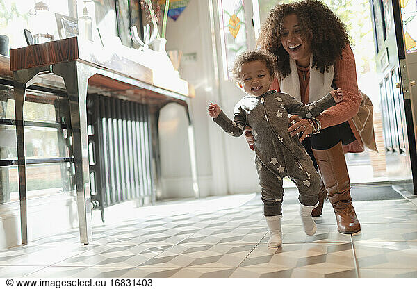 Happy mother helping baby daughter learn to walk at home
