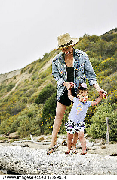 Happy mother holding hands of daughter while standing on fallen tree at beach