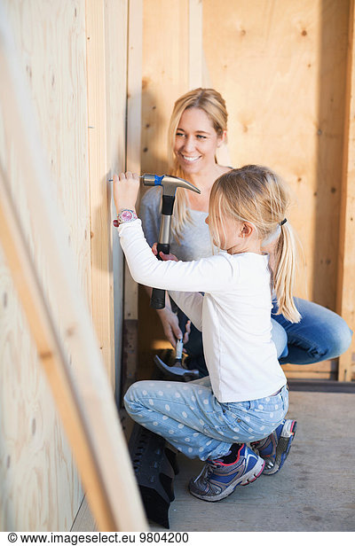 Happy mother looking at daughter hammering nail outside house being renovated