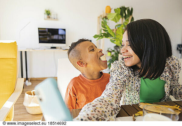 Happy mother taking selfie with son eating banana at home