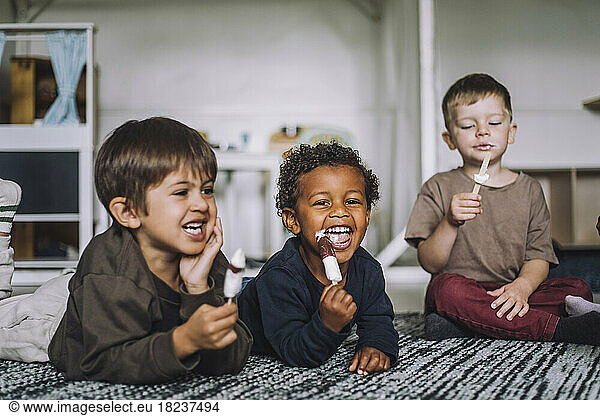 Happy multiracial male students eating ice creams on carpet at day care center