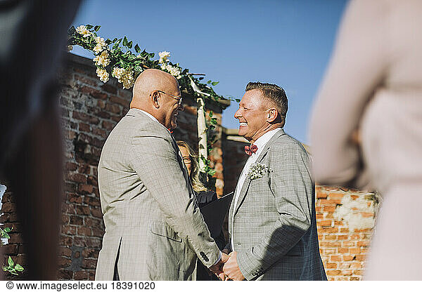 Happy newlywed gay couple holding hands by minister on wedding during sunny day
