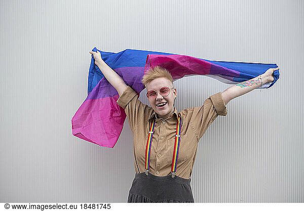 Happy non-binary person holding bisexual flag in front of white wall