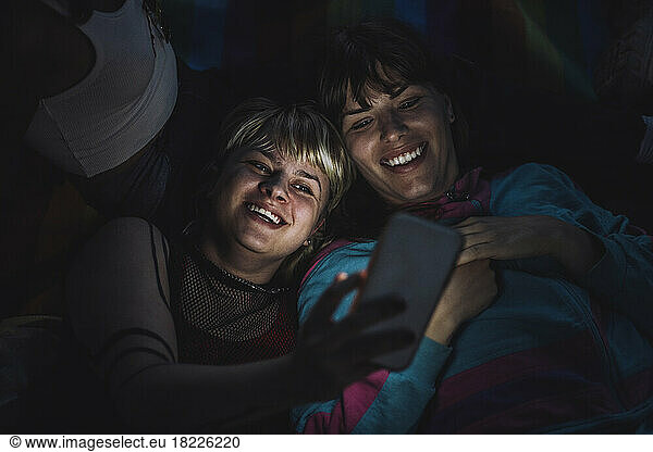 Happy non-binary person taking selfie with friend through mobile phone at night