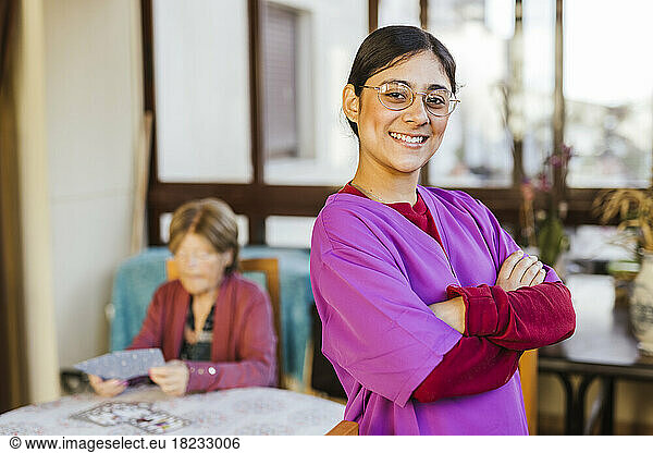 Happy nurse standing with senior woman sitting in background at home