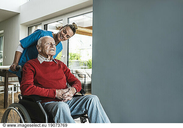 Happy nurse taking care of senior man sitting in wheelchair by wall at home