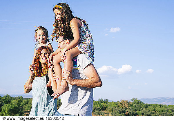 Happy parents carrying daughters on shoulders against sky during sunny day