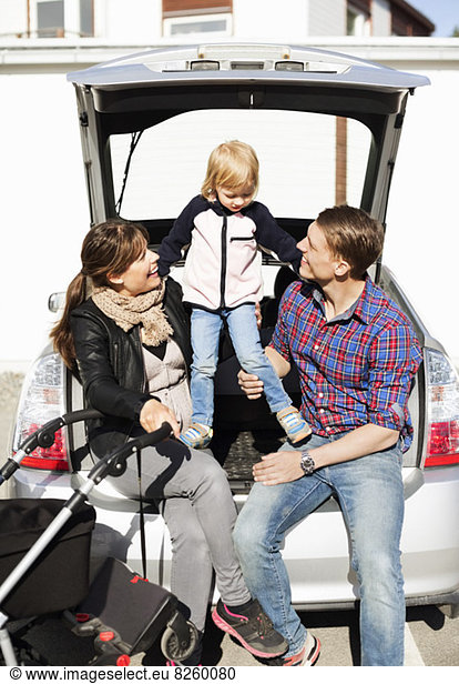Happy parents with daughter and baby carriage sitting in car trunk