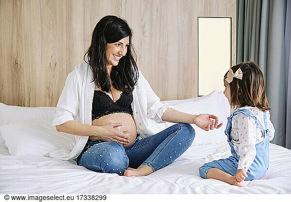 Happy pregnant mother looking at daughter while rubbing moisturizer on stomach