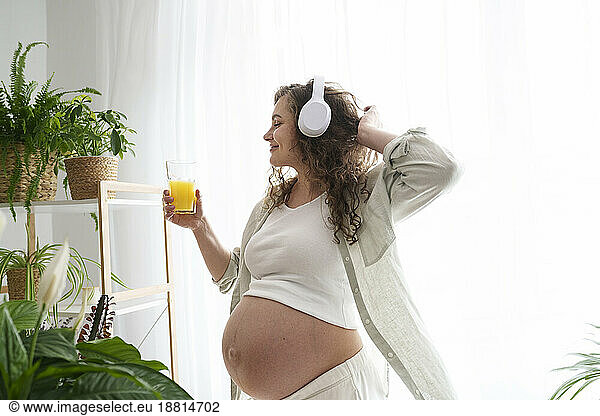 Happy pregnant woman enjoying listening music and having juice at home
