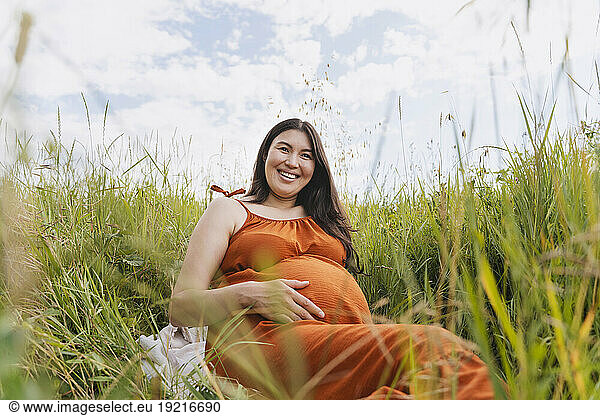 Happy pregnant woman sitting amidst grass under sky