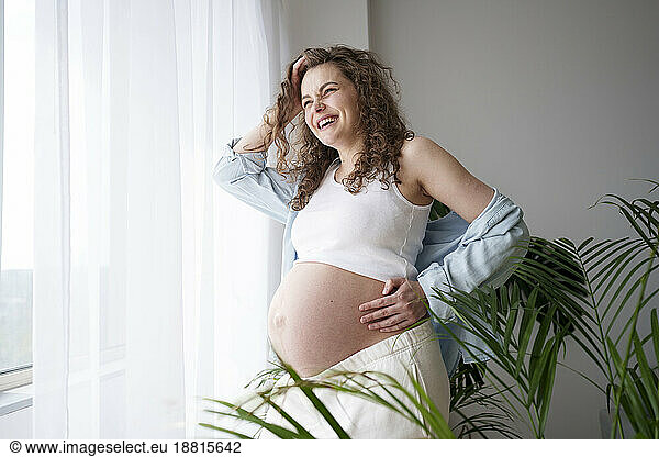 Happy pregnant woman with hand on hip standing near window at home