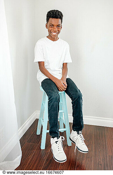 Happy preteen black boy in tshirt  jeans & sneakers on blue stool
