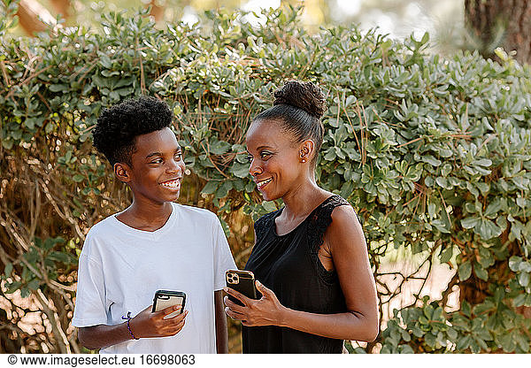 Happy preteen boy and mid-40's Black mom outdoors with iPhones