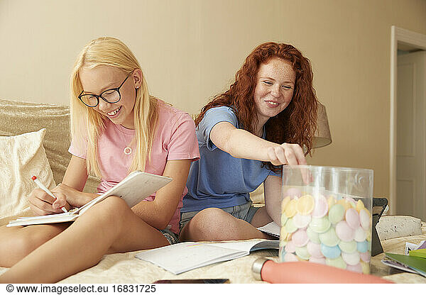 Happy preteen girl friends eating candy and studying on bed