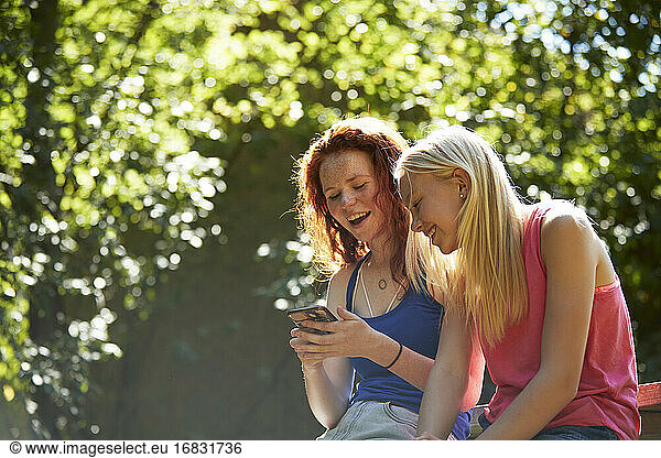 Happy preteen girl friends using smart phone below sunny trees