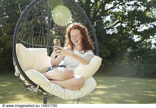 Happy preteen girl using smart phone in swing chair in sunny backyard