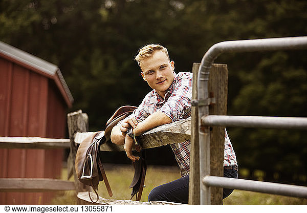 Happy rancher with saddle leaning on fence in farm