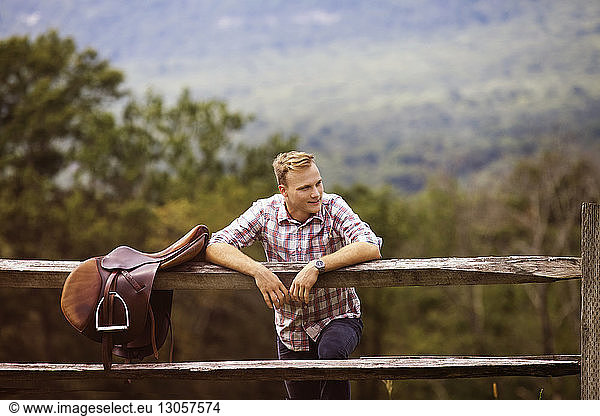 Happy rancher with saddle leaning on fence in farm