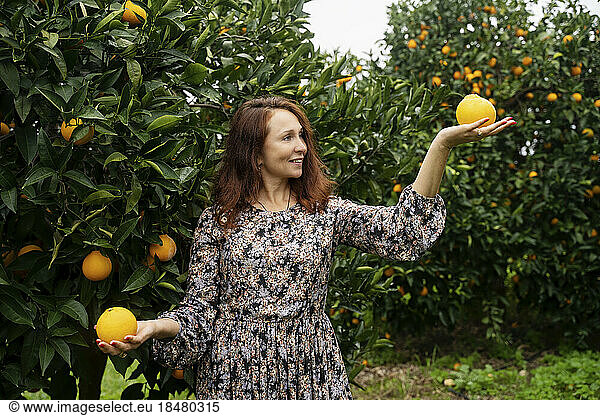 Happy redhead woman balancing oranges in front of trees
